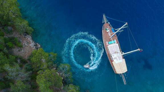 ein großes Segelboot, das in einer klaren, blauen Bucht vor Anker liegt. Ein Jet-Ski zieht Kreise neben dem Boot.