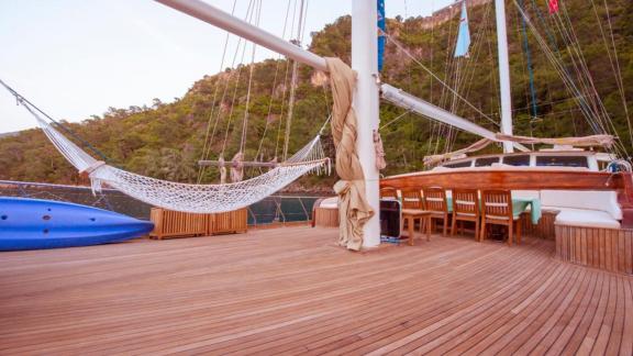 Deck of a large sailing boat with hammock and dining table in front of a picturesque coastline.