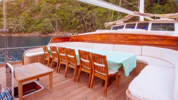 A dining area on the deck of a sailing boat with a long table and wooden chairs.