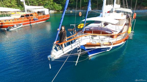 A woman stands on the bow of a gulet anchored in a calm bay.