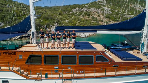 The professional crew of the SY Voyage gulet stands on deck with a scenic coastline in the background.