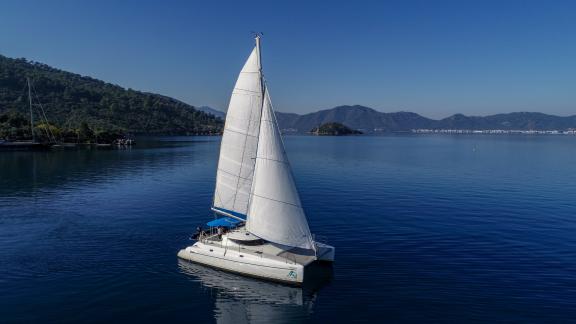 Andromeda catamaran glides on the blue sea with open sails, surrounded by mountain scenery.