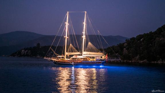 A beautifully illuminated gulet anchored at night in a tranquil bay.