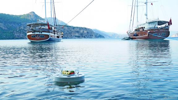 A floating table with drinks in front of two anchored yachts in a scenic bay.