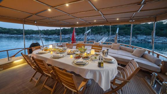 Stilvoll gedeckter Esstisch auf dem überdachten Deck der Gulet Torini, umgeben von ruhigem Wasser und grüner Küstenlands