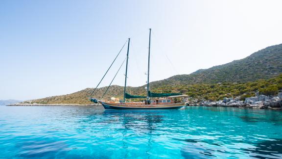 The gulet Ros Mare at anchor in turquoise waters, surrounded by hills and clear skies.