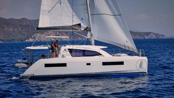 The catamaran Deniz3 sailing on the sea with sails up, with mountain views in the background.