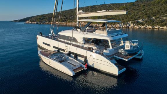 Der Lagoon Katamaran Adriatic Dragon ankert mit einem Beiboot an seiner Seite vor einer Küste.