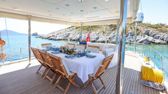The aft deck of Gazella features a dining table with a sea view and wooden chairs.