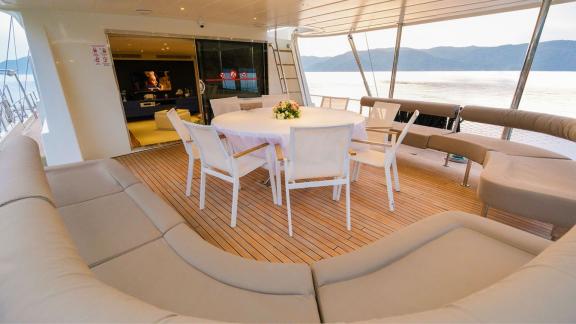 Dining area on the deck of the Gulet Vega with a round table and surrounding sofas, surrounded by calm water and mountai