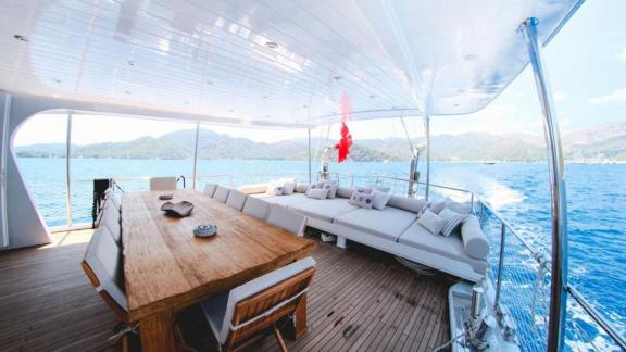 A dining table and a lounge area on the deck of a gulet with a view of the sea and the surrounding mountains.