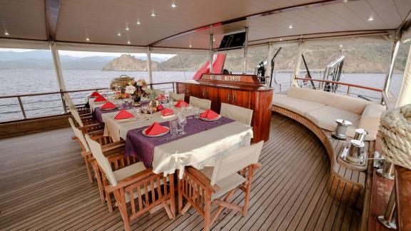 Dining area of a yacht with set table and chairs in front of a coastal landscape.