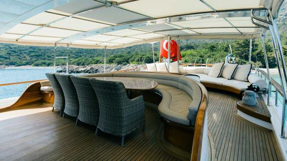 Elegant aft deck of the Gulet Caner 4 with covered seating area, dining table and view of the sea and the coast.