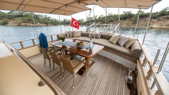 Cozy dining area on the deck of a gulet with table, chairs, and sofas in front of a scenic coastline.