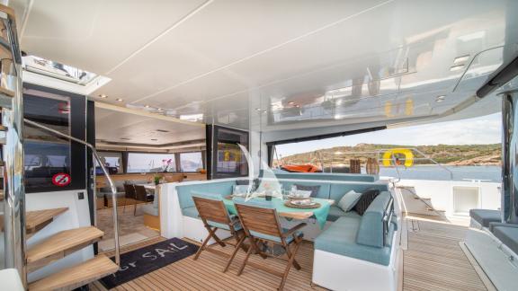 Covered outdoor deck with comfortable lounge, dining table, and stairs to the flybridge, overlooking the sea and coast.