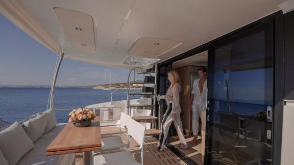 Two elegant guests stepping onto the spacious aft deck of a luxury catamaran, with stairs leading to the flybridge.