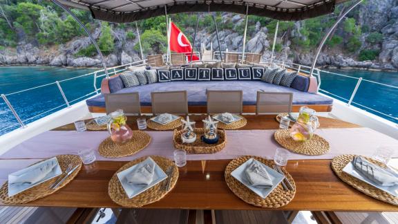 The picture shows a beautifully laid dining table and a luxurious lounge on the aft deck of the motorsailer Nautilus in