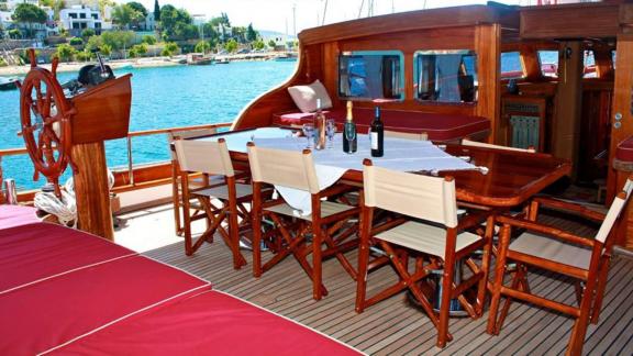 Set table, chairs, and red cushioned benches on the aft deck of Gulet Il Fratello with a view of the coast.