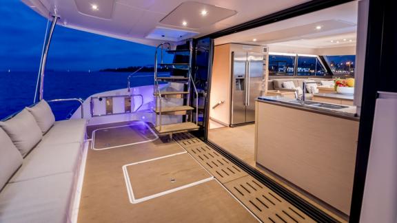 Illuminated aft deck of a modern catamaran at night, with a view into the luxurious open kitchen inside.