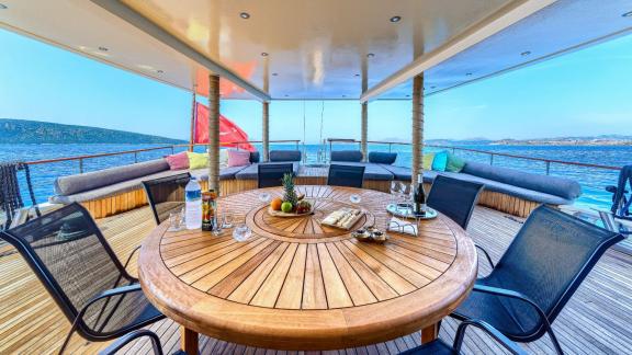 Covered dining area with round wooden table, chairs, sun loungers, Turkish flag and sea view.