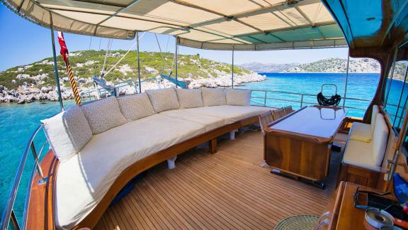 Spacious outdoor deck of Gulet Erden with cushioned seating and wooden table, surrounded by turquoise water.