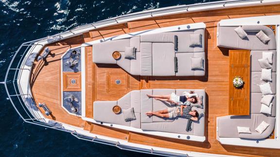 Two people relax on sunbeds on the spacious deck of motor yacht Nemesis with seating and a table.