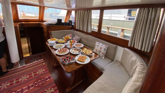 Уютная обеденная зона гулета Be Happy в гавани, богато уставленная различными блюдами.