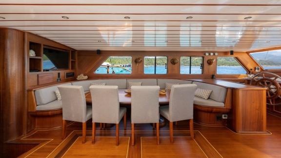 Wooden interior of a boat with dining table and six chairs, panoramic windows offer a view of the water.