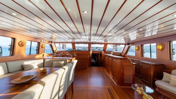 Wooden interior of a boat with dining table, sofa and steering wheel, panoramic windows offer a view of the water.