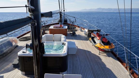 The deck of a sailing yacht with whirlpool, seating and jet ski, on a calm sea with mountains in the background.