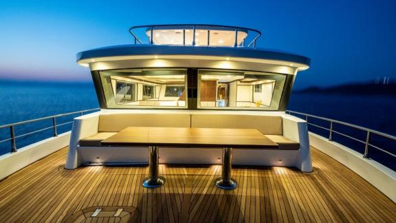 Wooden table and seating area on By CF trawler’s front deck under evening lights.