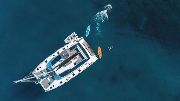 Catamaran seen from above, surrounded by kayaks and people swimming in clear water.