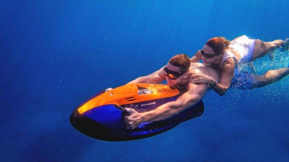 Man and woman riding an underwater scooter together in deep blue water.