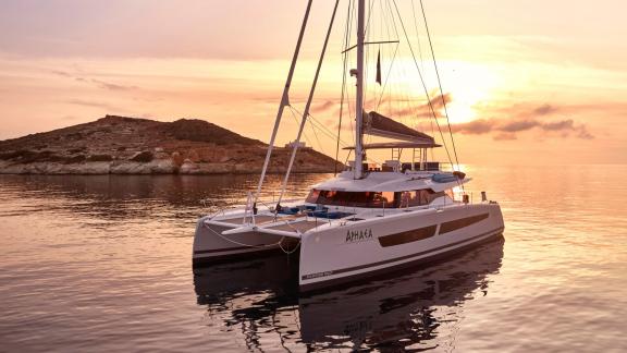 Der Katamaran Aphaea liegt ruhig vor Anker, während die Sonne hinter einer griechischen Insel untergeht.