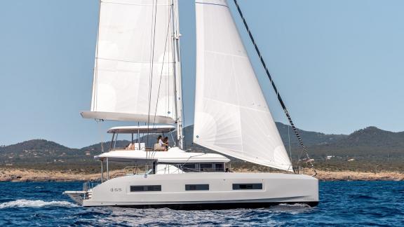 Lagoon 55 catamaran with full sails on calm seas, with a coastal backdrop of rolling hills.