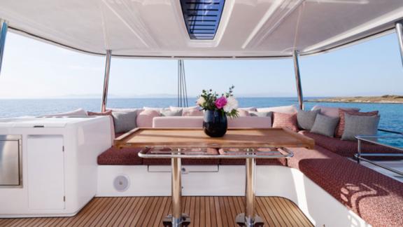 The flybridge of Dream catamaran features a wooden table, soft cushioned seats, and a spacious lounge.