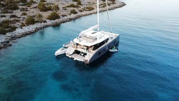 The Genny catamaran anchored in calm, turquoise waters near a secluded rocky shoreline.