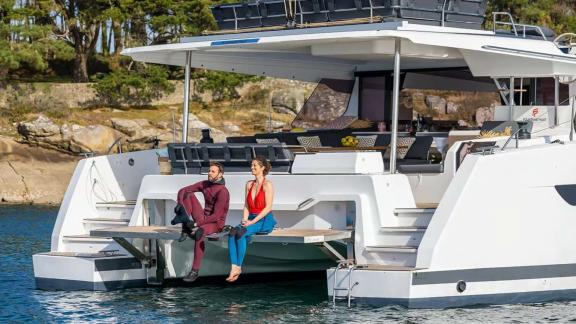 The catamaran Kebisiks is anchored as two guests sit in wetsuits on the aft platform.