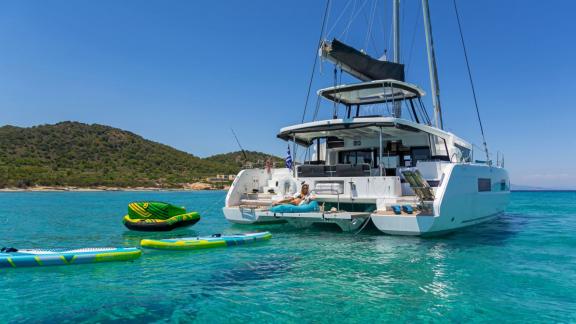 The yacht is anchored, a guest relaxes on the aft deck. Paddleboards and water toys are in the water.