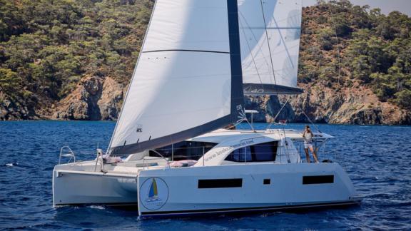 Pata catamaran sailing on the sea with rocky cliffs and trees in the background.