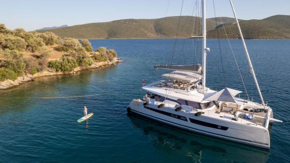 The catamaran Serenissima is anchored near a wooded Mediterranean island with a paddleboard in the water.