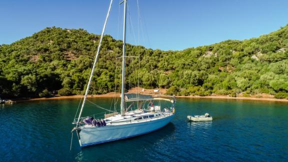Парусная яхта Conformista стоит на якоре в тихой бухте, окруженной зеленью.