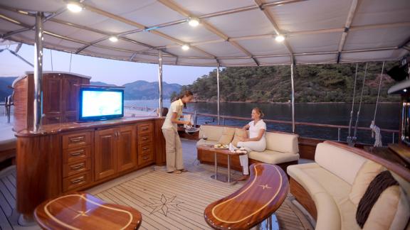 Lounge area with wooden furniture on yacht deck, crew serving guests by calm coast