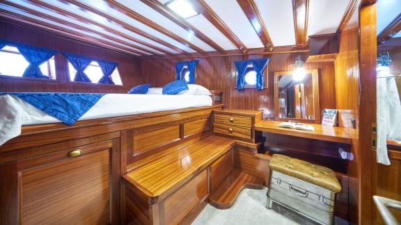 Stylish cabin with a raised double bed, wooden walls, blue curtains and a small desk with mirror on a gulet