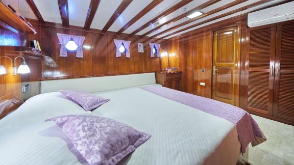 Cabin with wooden walls, double bed, purple bed linen, windows with curtains and built-in wardrobe.