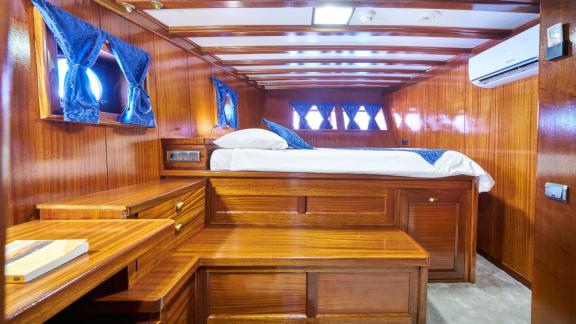 Bedroom cabin on a sailing yacht with wooden panelling, double bed, blue curtains and a small desk.