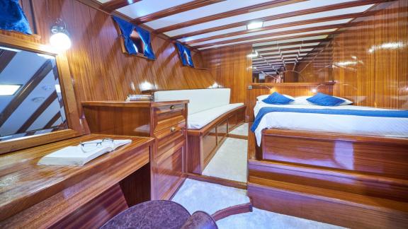 Wood-panelled cabin on a sailing yacht with a double bed, white and blue bed linen, a bench and a desk