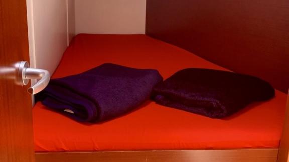 The cabin of the Deneb sailing yacht features a single bed with a red cover and dark blankets.