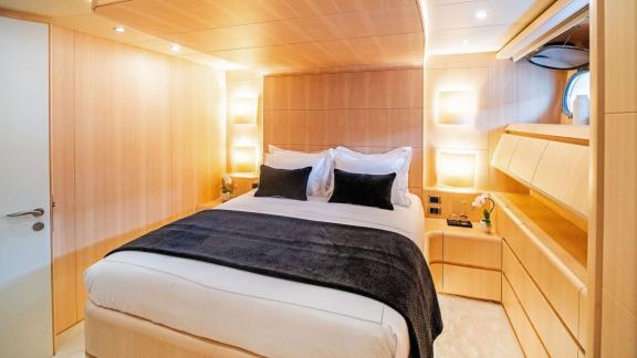 The cabin features a double bed, light wood furniture, wall lights and a round porthole window.