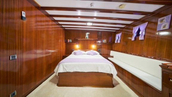 Wood-panelled bedroom on a sailing yacht with a double bed, white and purple duvet covers and a comfortable bench.